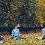 Pretty young women are relaxing sitting in lotus position on yoga mats in park and breathing fresh air resting after outdoor group practice. Meditation and nature concept.